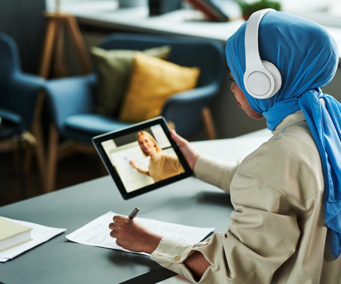 Young Muslim woman in hijab and headphones making notes during online lesson