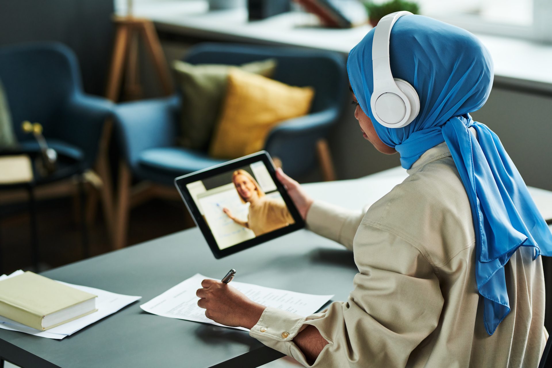 Young Muslim woman in hijab and headphones making notes during online lesson