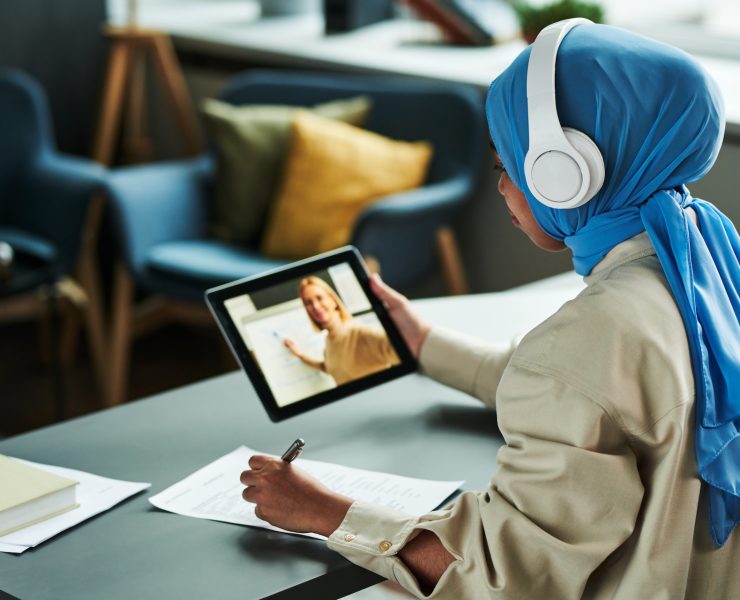 Young Muslim woman in hijab and headphones making notes during online lesson