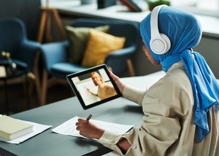 Young Muslim woman in hijab and headphones making notes during online lesson